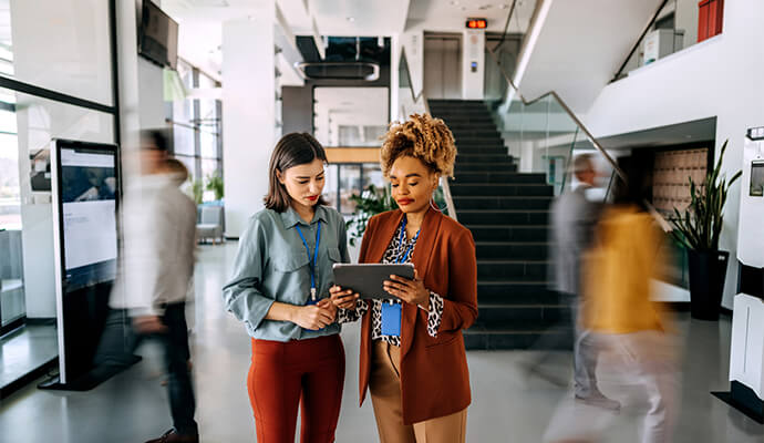 two-women-looking-at-tablet-in-office-open-space-blurred-people-motion-bdo